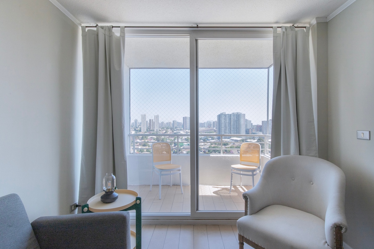 A living area is shown with large sliding glass doors opening to a balcony, revealing a city skyline. Two light-colored chairs are positioned outside on the balcony, and a cozy armchair and small table are located indoors, enhancing the space's bright ambiance.