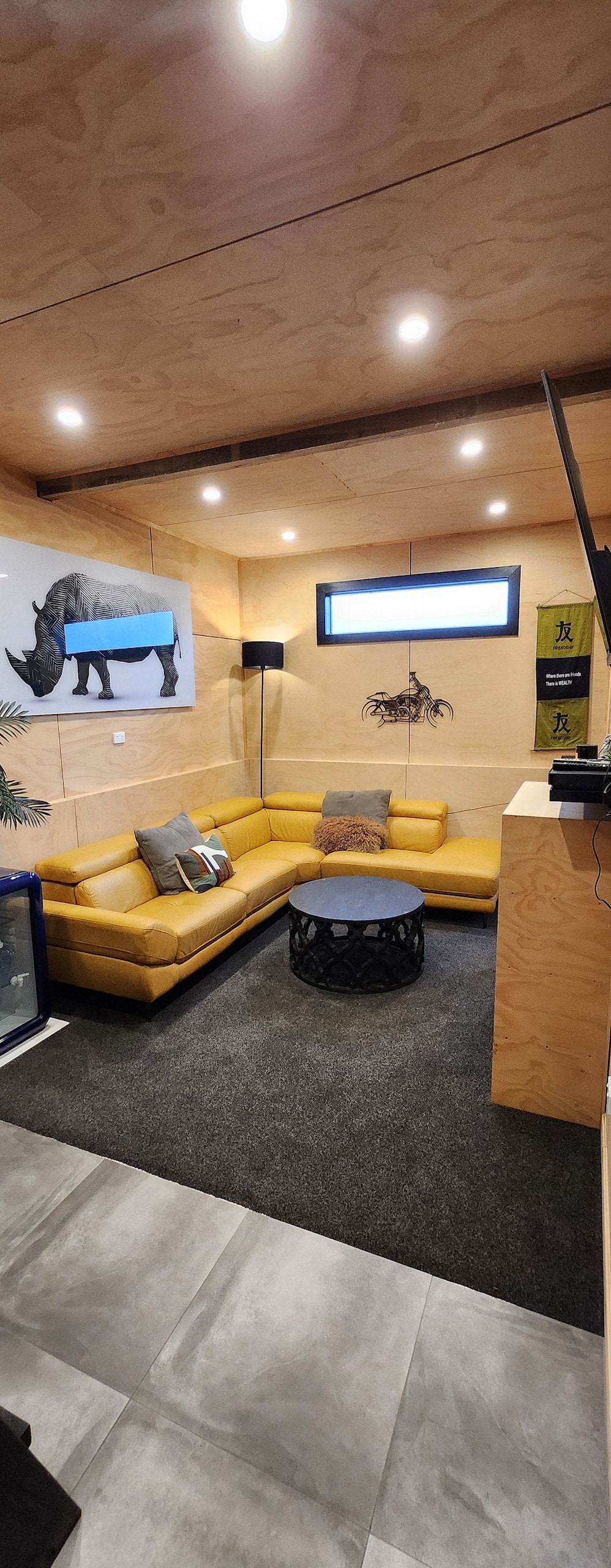 A cozy living area features a yellow sectional sofa with decorative pillows, surrounding a round black coffee table. Soft lighting illuminates the warm wood paneled walls, while a large window offers natural light. A modern art piece and a framed photo adorn the wall.