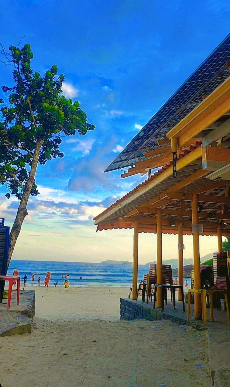 A sandy beach is visible, framed by a vibrant blue sky with scattered clouds. A beachfront structure with a wooden roof provides shade, featuring tables and chairs arranged on a terrace. The ocean is in view, with distant beachgoers enjoying the shoreline.