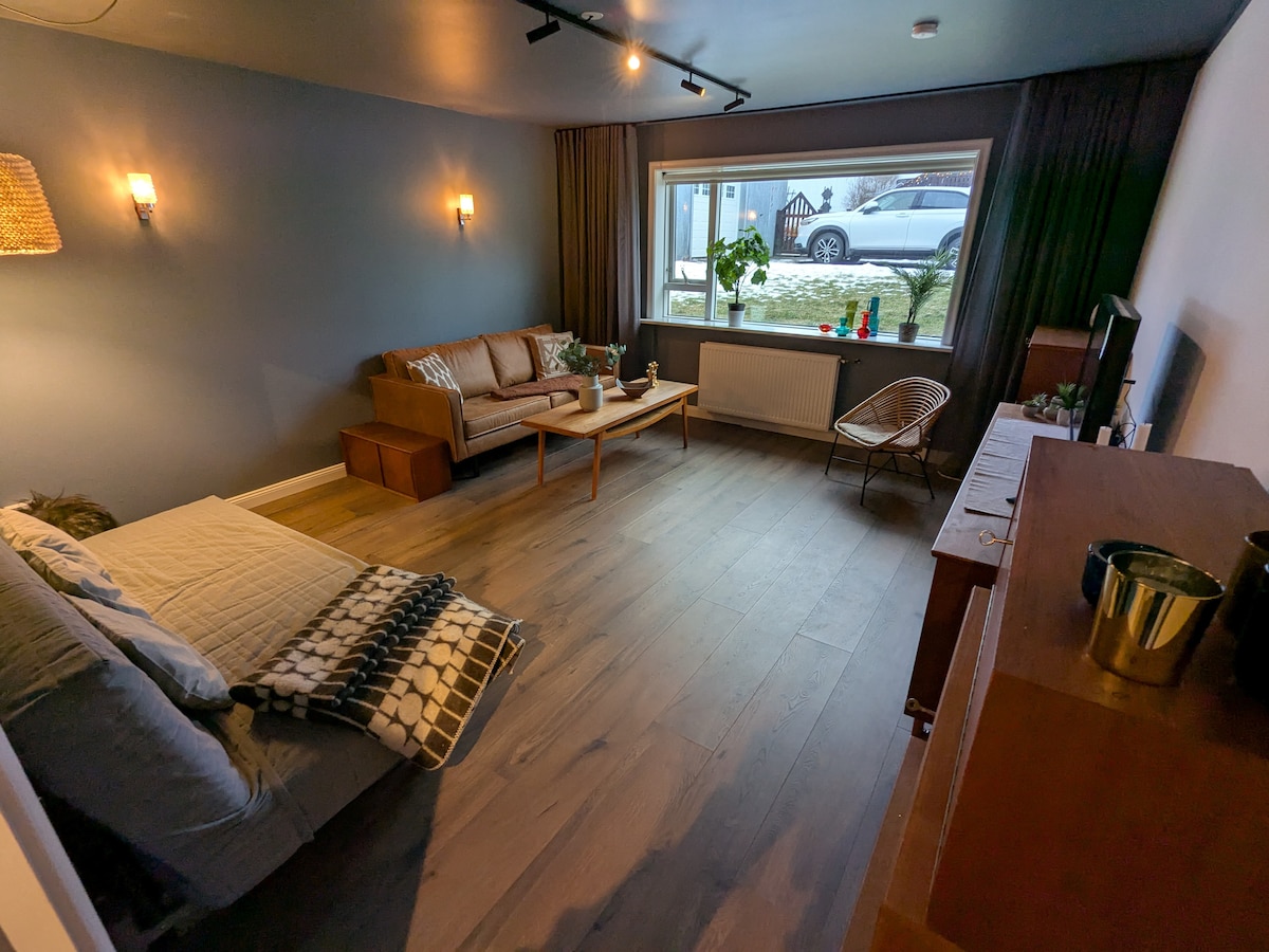 A spacious living room is featured, showcasing a comfortable sofa and an additional sofa-bed. Natural light streams in through a large window, revealing outdoor views. A wooden table rests in front of the sofa, and decorative plants add a touch of greenery.