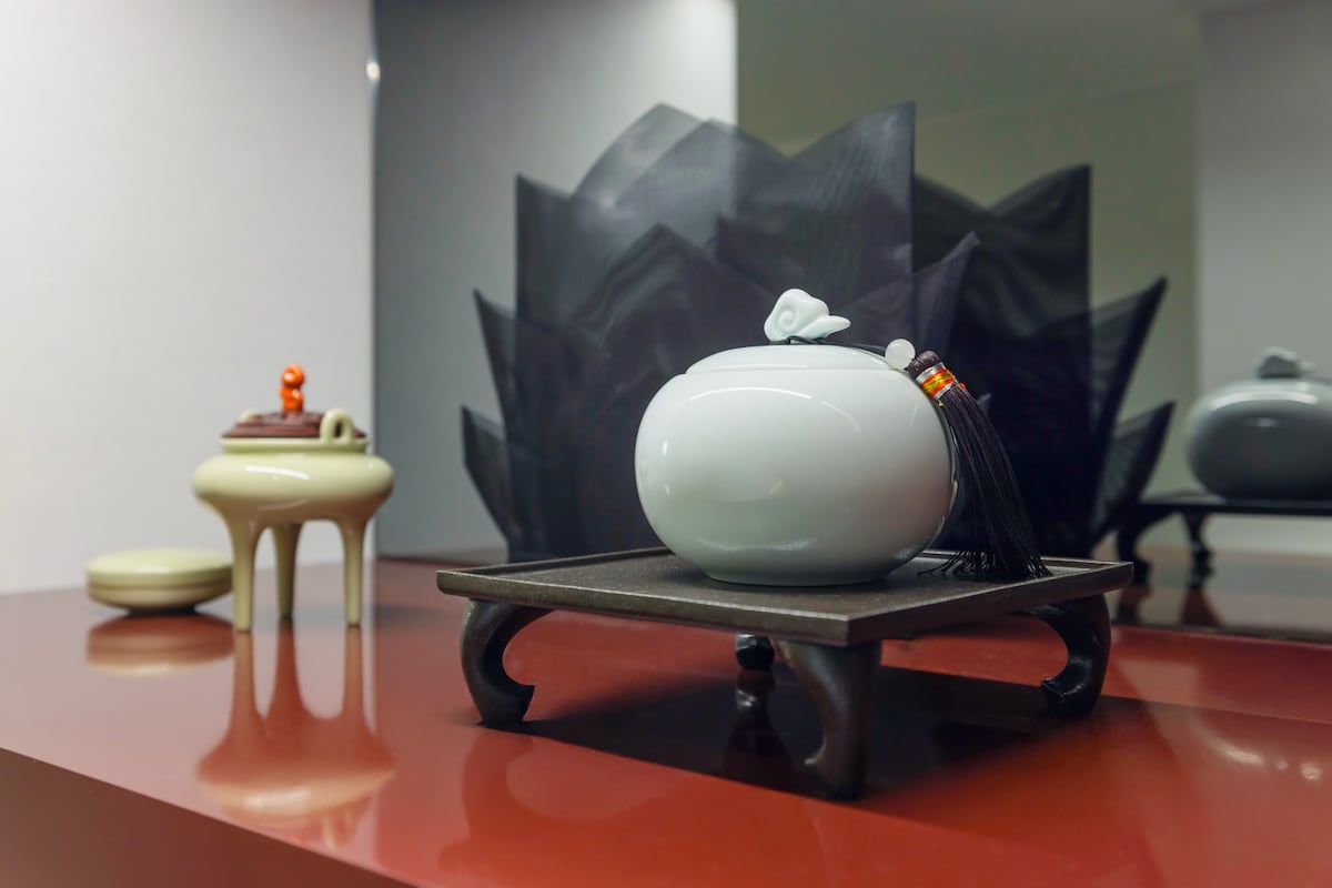 A decorative ceramic piece is displayed on a dark wooden stand, showcasing an elegant design. Next to it, a small, rounded stool features a light-colored surface. A sculptural backdrop adds depth, with a design resembling layered petals, contributing to the serene decor.