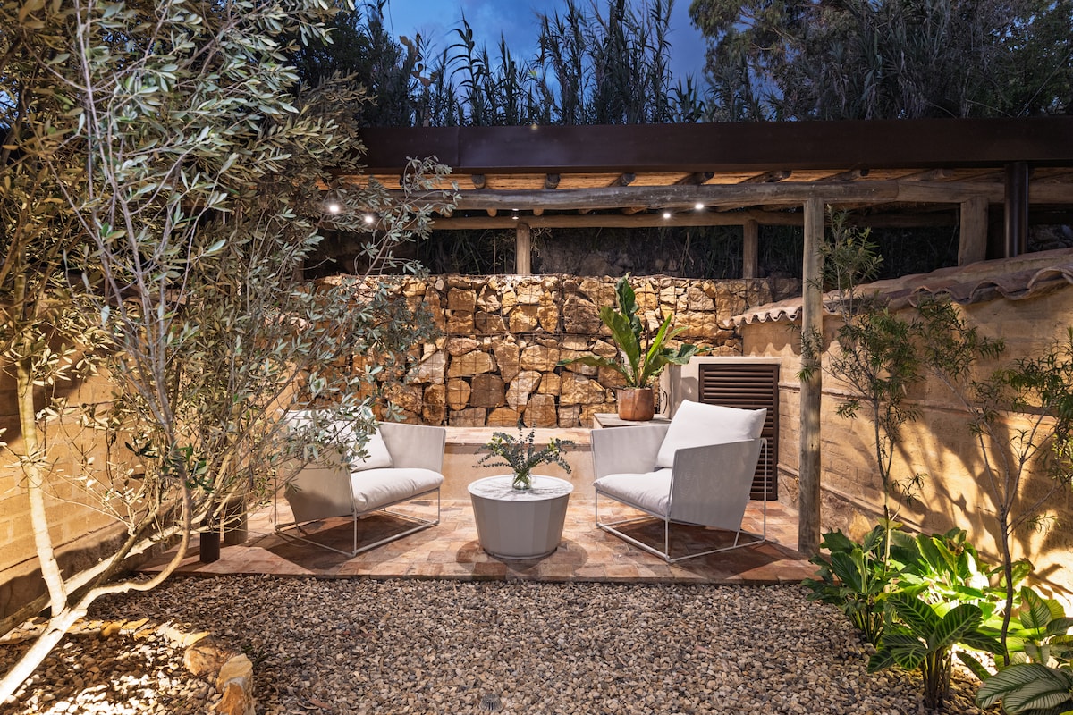 An outdoor seating area is presented, featuring two white armchairs positioned around a round table. Lush greenery is integrated into the space, with stone walls providing privacy. Soft lighting enhances the serene atmosphere, and natural elements like olive trees are visible in the background.
