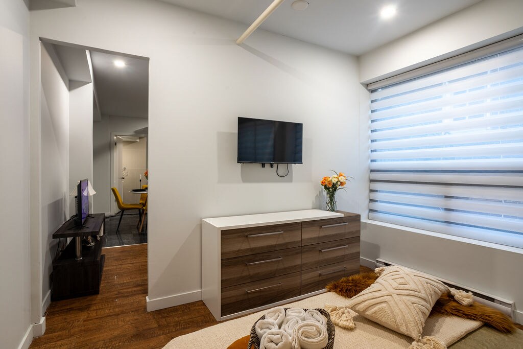 A welcoming bedroom features a spacious bed adorned with neutral linens and a cozy throw. A dresser with a modern design is present beneath the wall-mounted TV, while a small vase of fresh flowers adds a touch of color. Light filters through the window with sheer blinds.