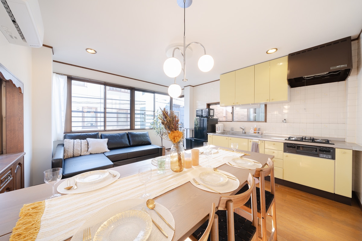 A light-filled kitchen and dining area is displayed, featuring a spacious dining table set for mealtime. A sofa is positioned near large windows, allowing natural light to brighten the space. The kitchen includes yellow cabinetry and essential appliances, creating a functional atmosphere for cooking and dining.