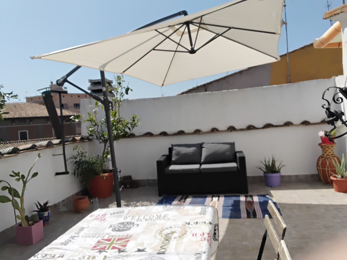 A spacious terrace is adorned with a comfortable sofa under a large umbrella. A patterned table is set nearby, while potted plants add greenery to the area. The surrounding walls are white, enhancing the outdoor seating space for relaxation.