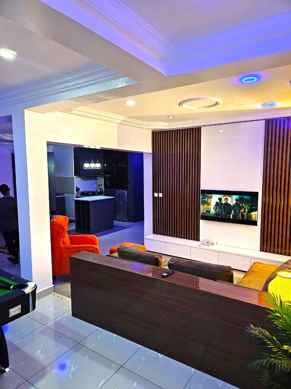 A modern living area is showcased with a stylish sofa and a large flat-screen TV mounted on a feature wall. Soft lighting creates a calming ambiance. The open layout leads to a kitchen area in the background, visible through a spacious entrance.