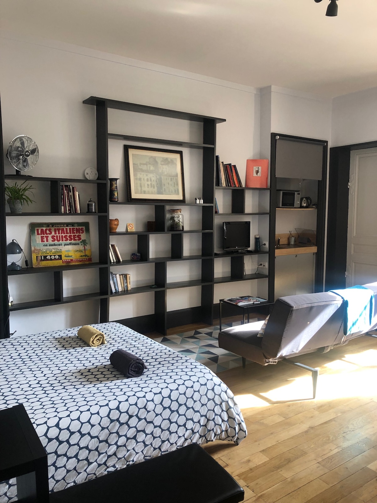 A spacious living area features a patterned bedspread and a cozy seating arrangement, complemented by a wall-mounted shelf displaying books and decor items. Light filters through the window, illuminating the wooden floor and contemporary furnishings.