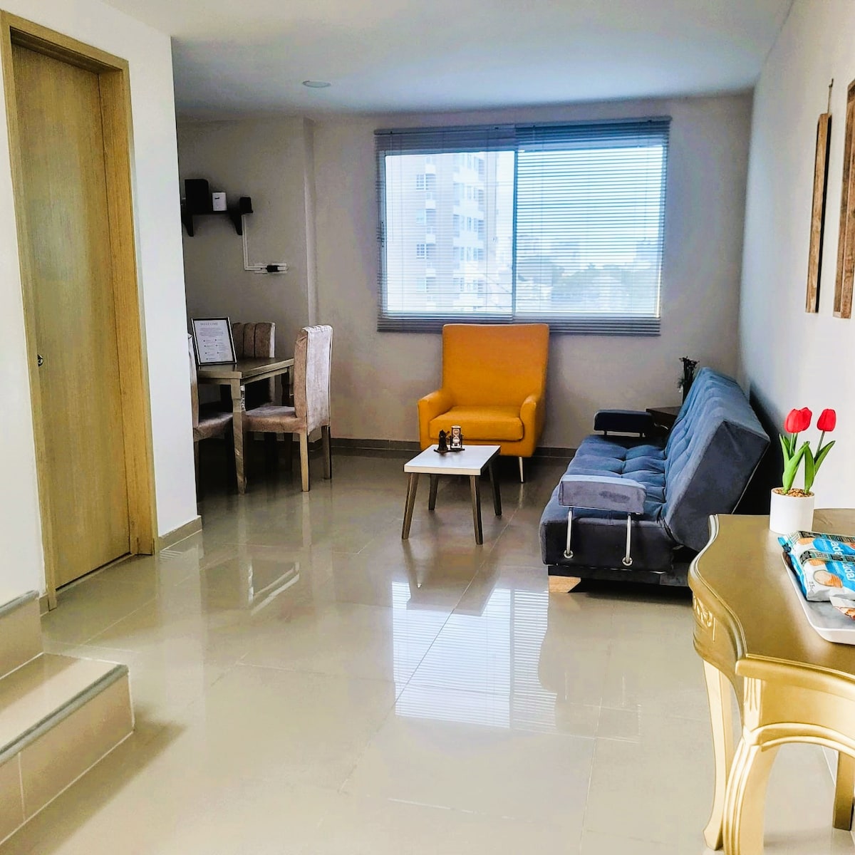 A living area is presented with a modern sofa bed, a small coffee table, and a bright yellow accent chair. A dining table with upholstered chairs is positioned nearby. Large windows allow natural light to fill the space, enhancing the neutral tones of the decor.
