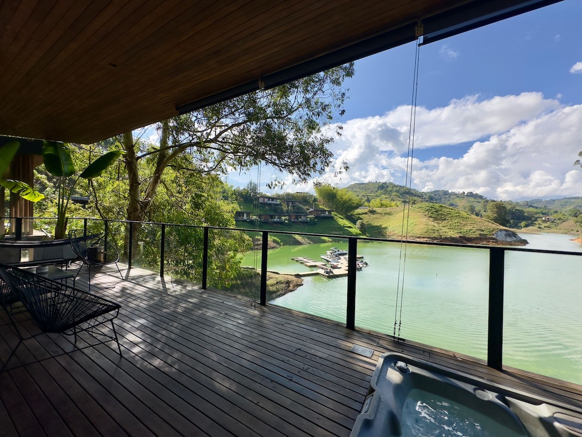 An outdoor terrace features a jacuzzi with views of a serene lake and surrounding hills. Lush greenery frames the scene, and a wooden deck extends into the space. The calm water reflects the sky and is punctuated by a private dock in the distance.