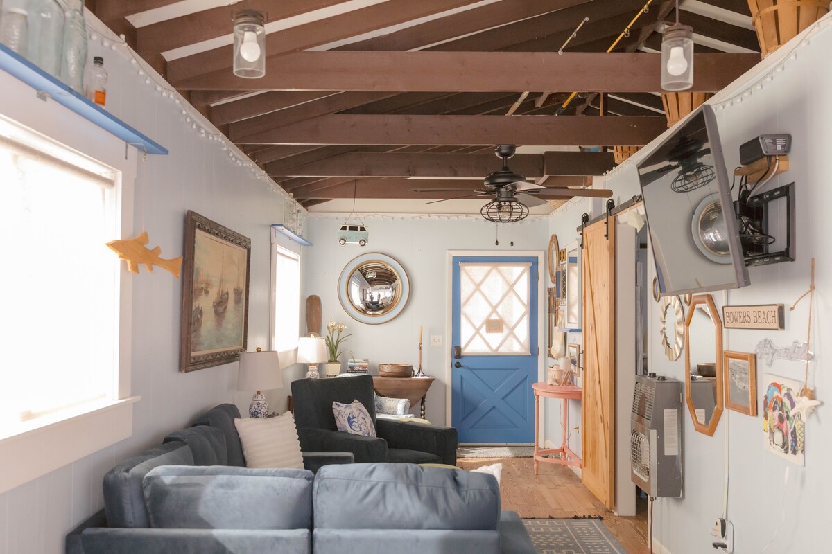 A cozy living area features a blue couch complemented by decorative pillows. A ceiling fan hangs from the vaulted ceiling, which showcases exposed wooden beams. A bright blue door adds a pop of color, while natural light filters through the two windows adorned with sheer curtains.