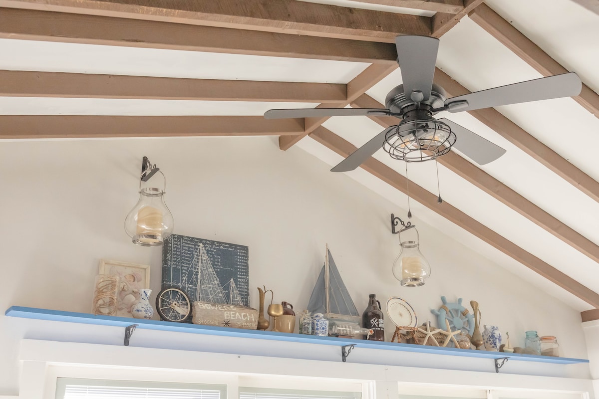 A ceiling fan with sleek blades hangs from a vaulted ceiling adorned with exposed beams. A light blue shelf displays various beach-themed decor, including lanterns, model sailboats, and nautical figurines, contributing to a coastal aesthetic.