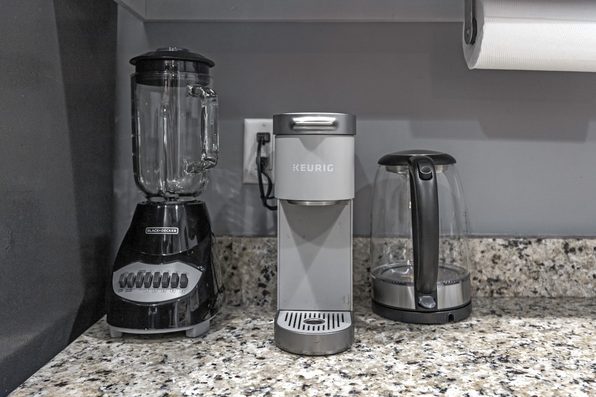 A modern kitchen countertop is displayed, featuring a black blender, a silver Keurig coffee maker, and a glass electric kettle. The granite surface is complemented by a smooth, gray backsplash.
