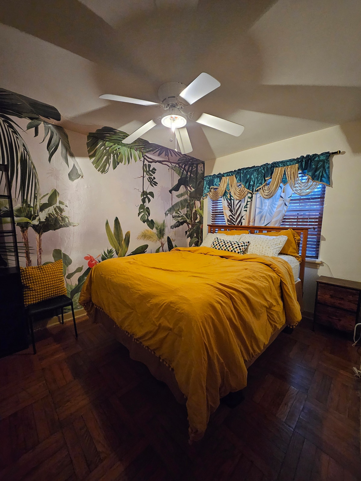 A cozy bedroom features a spacious bed dressed in a bright yellow coverlet. Tropical-themed wallpaper adorned with lush greenery serves as a backdrop. A ceiling fan provides airflow, while window treatments with teal accents complement the warm tones. A small chair adds to the room's functionality.