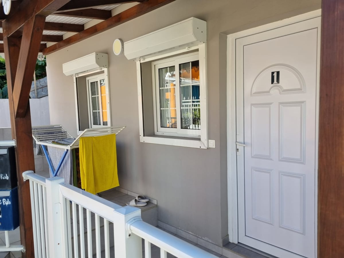 A welcoming entrance is featured with a white door numbered '1,' accompanied by two windows with white frames. A clothes drying rack holds vibrant yellow and blue items, positioned beside a railing that leads to the entrance.