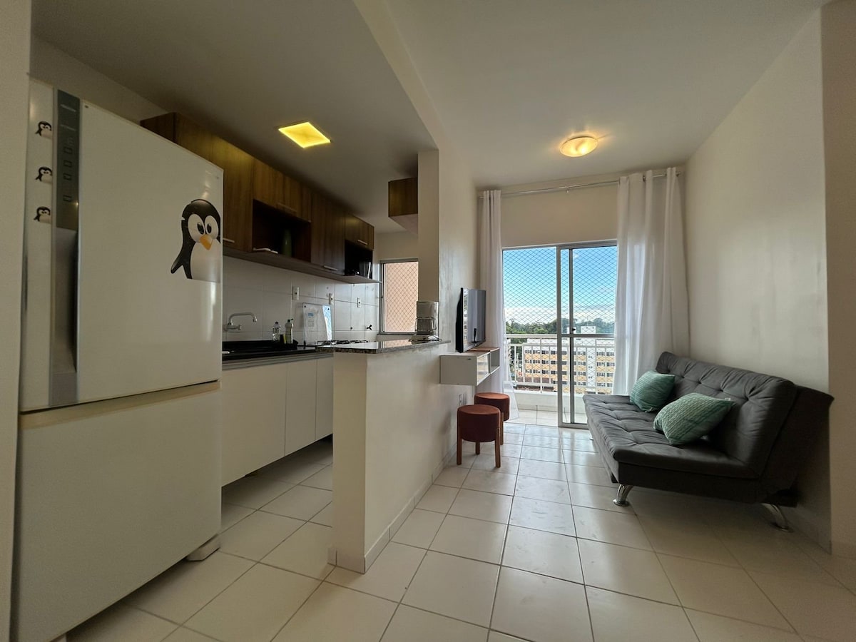 An open-concept kitchen and living area is presented, featuring a sleek refrigerator and a spacious countertop. A comfortable sofa bed sits next to a window, which allows natural light to brighten the space. The decor includes two green cushions and a small accent chair.