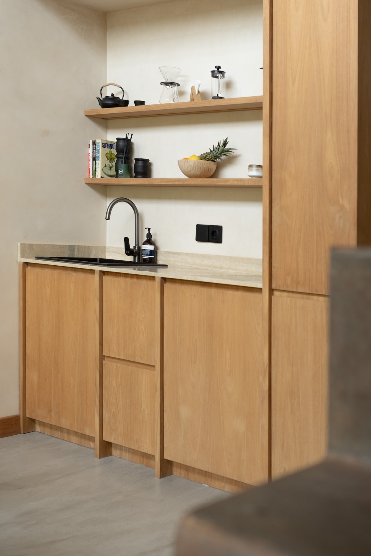 A minimalist kitchenette features wooden cabinetry with a countertop sink. Shelves above display cooking tools, books, and decorative items. The area is well-lit, showcasing a functional design that blends simplicity and warmth.