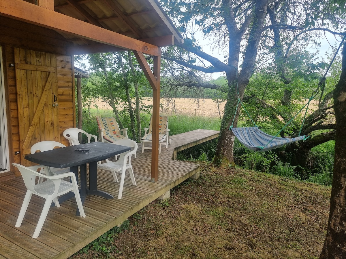 An outdoor terrace is presented with a dark table and four white chairs, surrounded by lush greenery. A comfortable hammock is seen hanging from a tree nearby, with a wooden walkway extending toward a small stream, offering a peaceful natural setting.