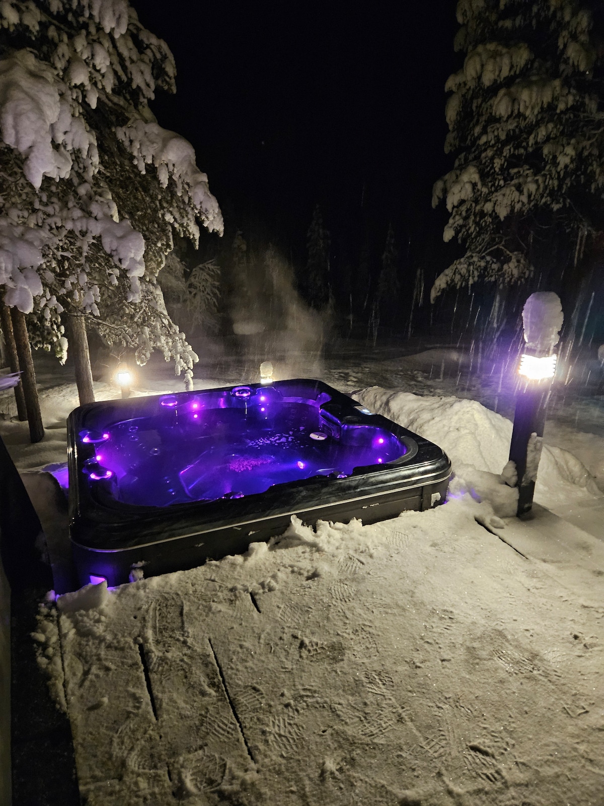 An outdoor hot tub surrounded by snow-covered ground and pine trees is illuminated with purple lighting. Steam rises from the water, creating a cozy atmosphere. The surrounding area is dimly lit, enhancing the serene winter night setting.