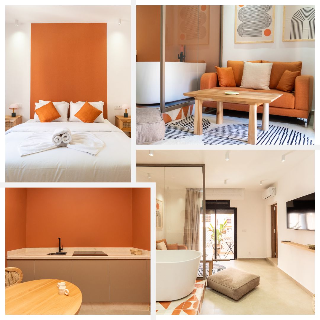 A spacious bedroom features a warm terracotta accent wall behind a large bed adorned with neatly arranged towels. An inviting sitting area with an orange sofa and a wooden coffee table is included. A modern kitchenette with minimalist cabinetry and a stylish bath area with a freestanding tub are also visible.