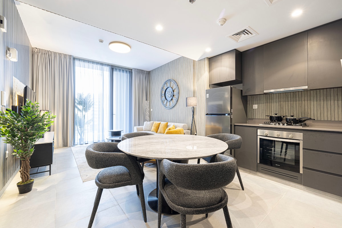 A modern kitchen and dining area features a round table surrounded by six chairs. Sleek cabinets and appliances are visible, including a refrigerator and an oven. Natural light enters through large windows, illuminating the stylish décor including a potted plant in the corner.