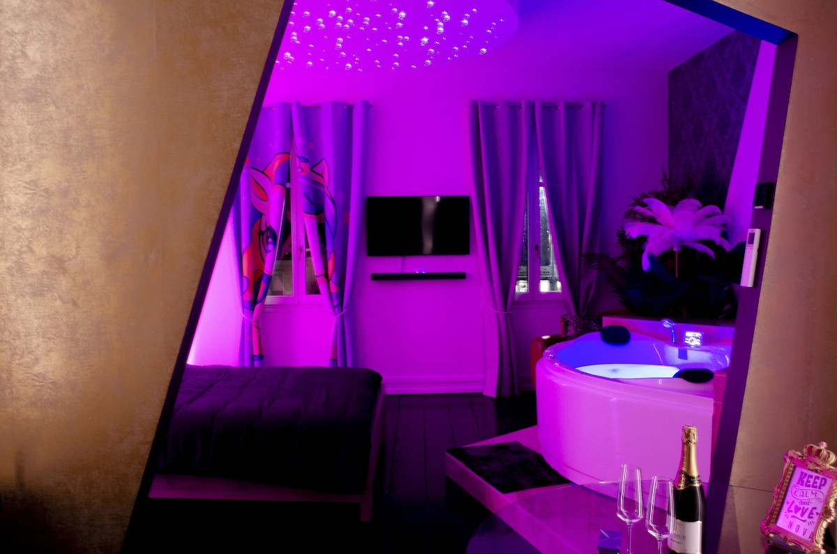 A modern loft-style room is illuminated in purple hues, featuring a spacious bed and a unique jacuzzi with multiple jets. Large windows are dressed with dark curtains, and a television is mounted on the wall. Decorative plants add a touch of greenery to the space.