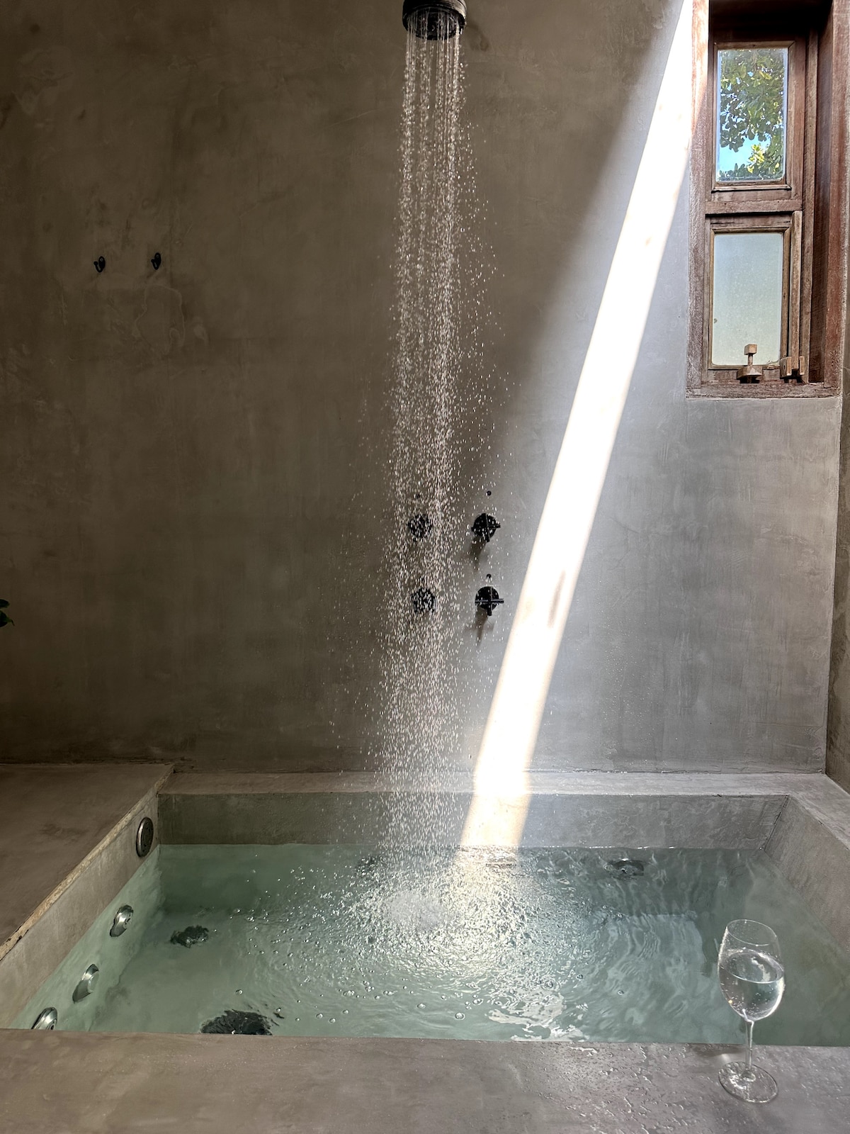 A serene bathroom scene features a jacuzzi with bubbling water, illuminated by a shaft of natural light. An elegant showerhead delivers a cascading flow of water overhead. A glass sits nearby, reflecting a touch of sophistication in the minimalist, textured concrete setting.
