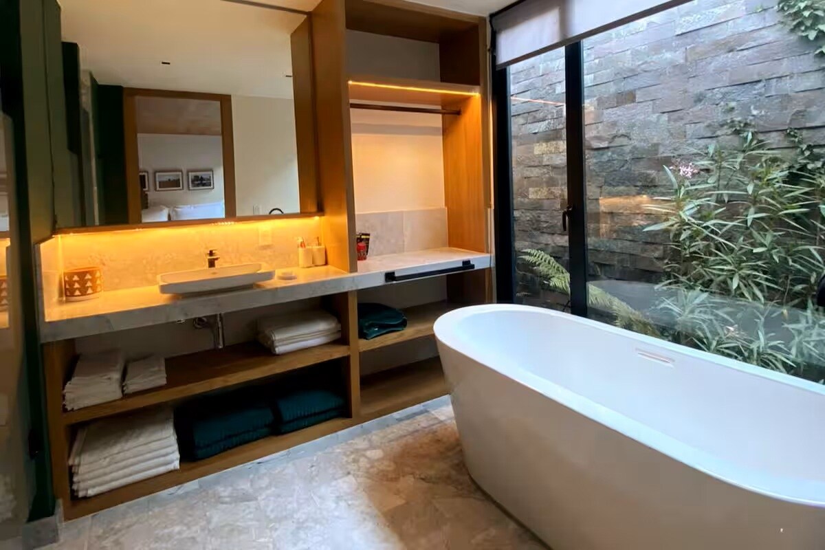 A spacious bathroom features a freestanding bathtub alongside a large window overlooking a serene garden. Natural light illuminates the room, highlighting the stone walls and wooden accents. Shelves display neatly stacked towels and toiletries, creating a functional and calming retreat.