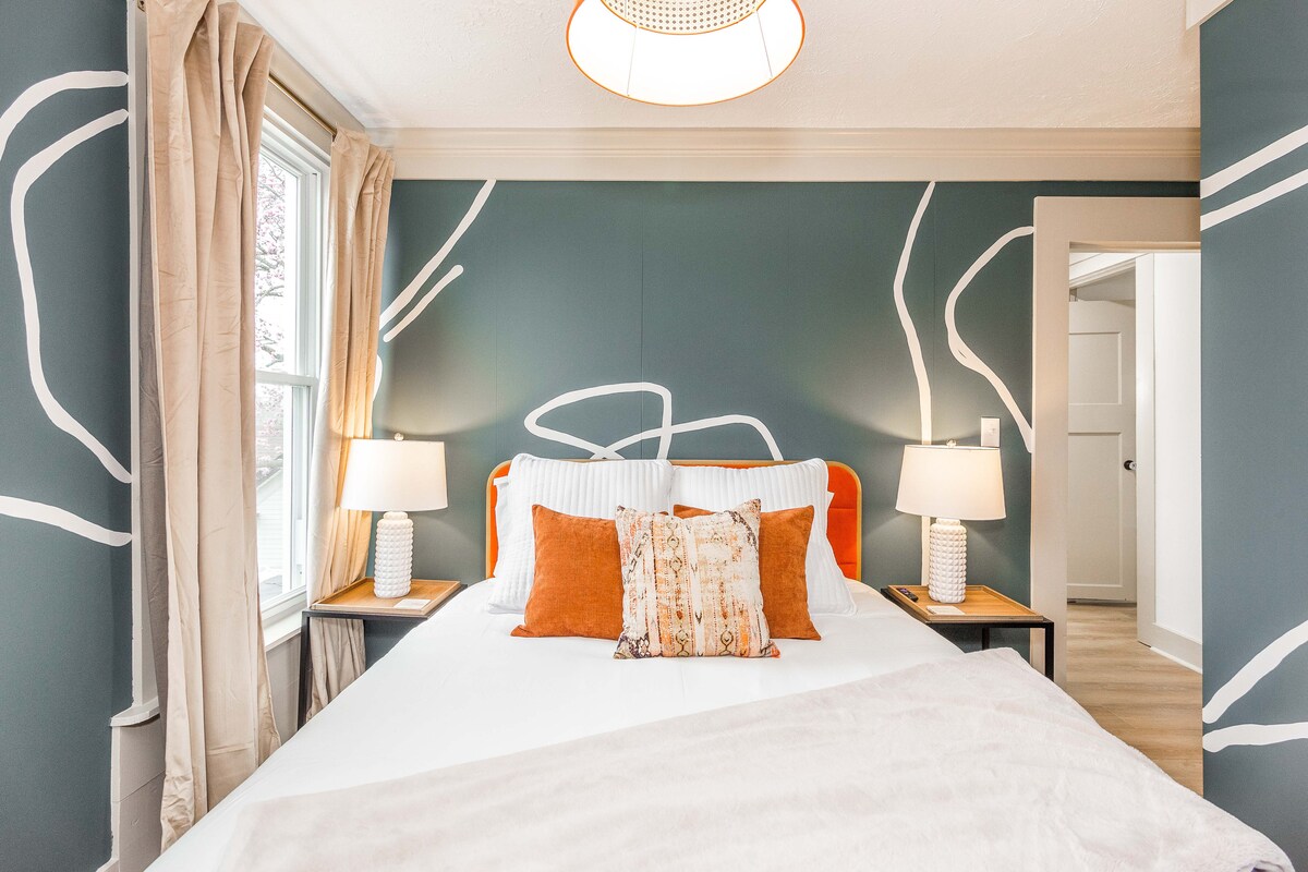 Модерна спалня със сини стени и артистични дизайни от бяла линия. Оранжевата кадифена рамка на леглото изскача на хладните стени, докато съчетаването на нощни лампи и слоести възглавници създава стилно, съвременно място за отдих.