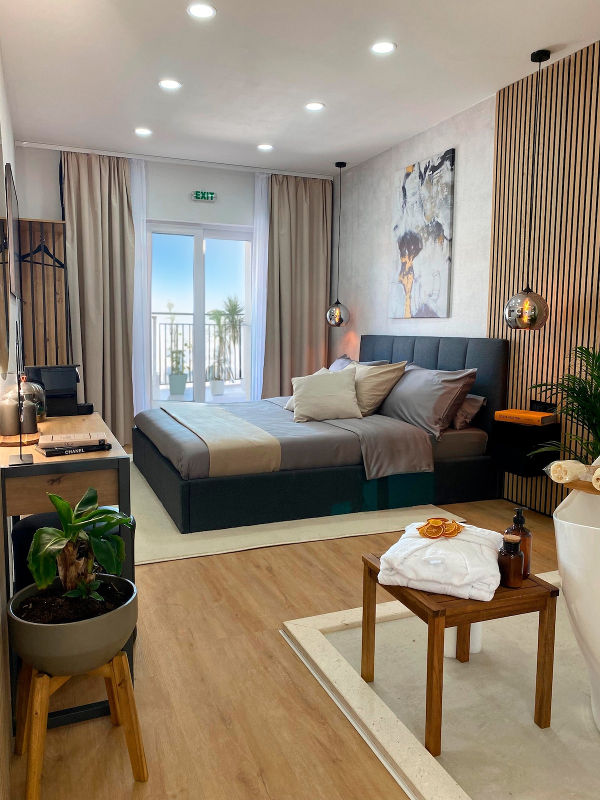 A comfortable room features a spacious bed with neutral linens and accent pillows. Natural light fills the space, highlighting a small table with a plant and toiletries. A stylish artwork is displayed on the wall, complementing the modern decor. The balcony view is glimpsed through glass doors.