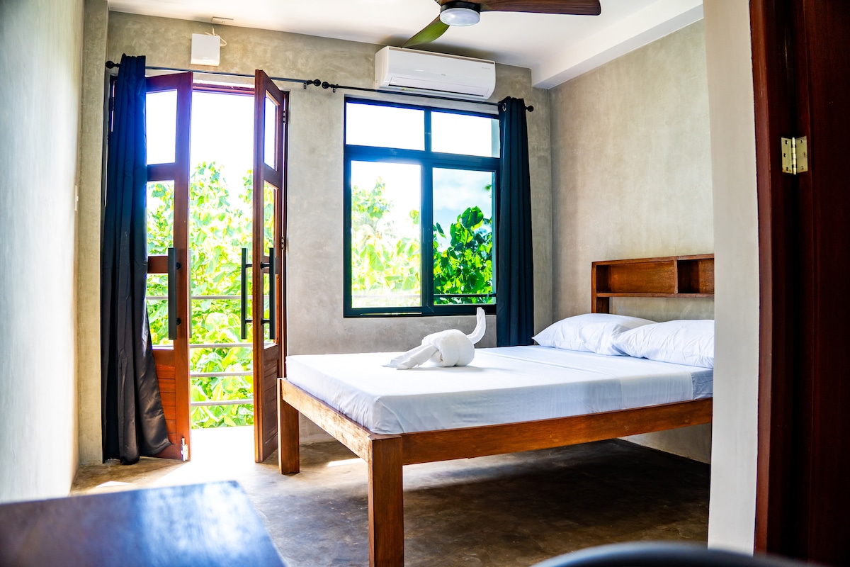 A bright sleeping area features a spacious bed with fresh linens. Natural light enters through a large window and double doors, which open to a balcony overlooking green trees. A modern ceiling fan is positioned above, contributing to the airy environment.