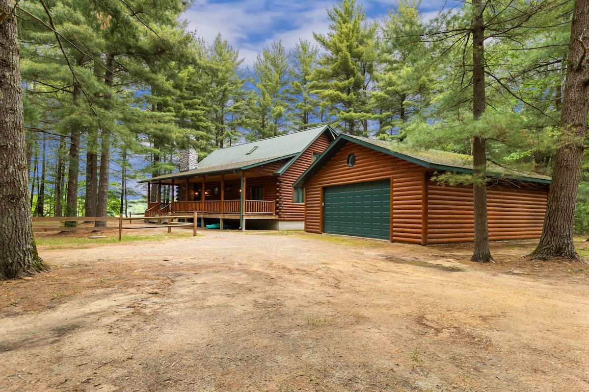 A rustic cabin is nestled among tall trees, featuring a welcoming wooden porch that wraps around the front. A large garage is attached to the side, and a gravel path leads to the entrance, enhancing the serene outdoor setting.