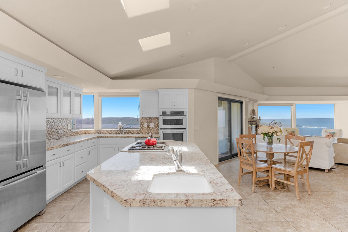 A spacious kitchen area features modern stainless steel appliances and a large island with a marbled countertop. Natural light pours in through skylights, illuminating a dining table set for four, while panoramic ocean views are visible through sliding glass doors.
