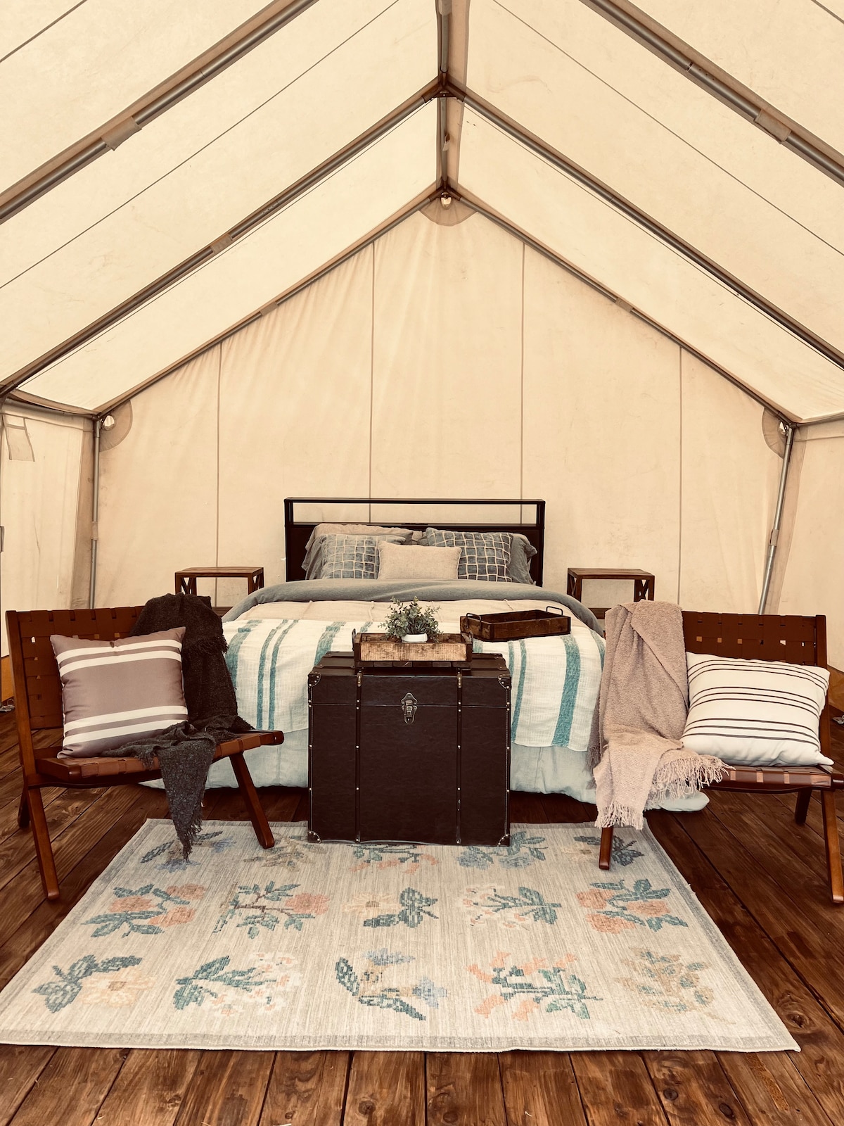 An inviting tent interior features a plush queen-size bed dressed in soft linens with a patterned throw draped over it. Two wooden chairs sit beside the bed, accompanied by a rustic trunk. A textured area rug adds warmth to the wooden floor.