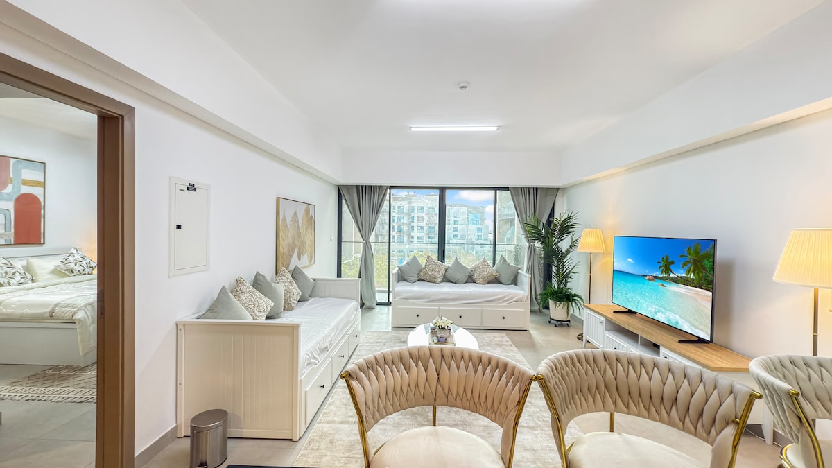 A spacious living area is presented, featuring a comfortable seating arrangement with light-colored chairs. A large television displays a beach scene, while large windows allow natural light to fill the room. Decorative pillows and cozy throws add a welcoming touch to the space.