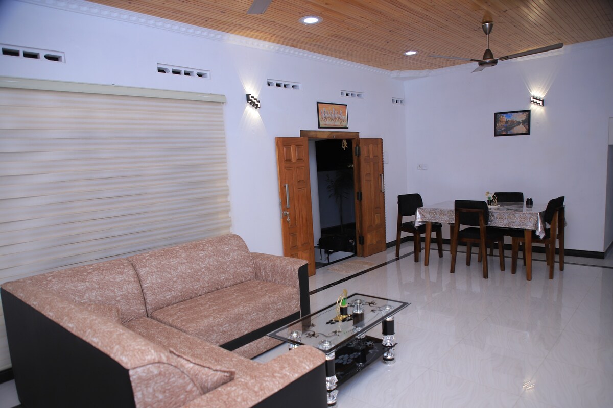 A spacious living area is presented, featuring a light-colored couch paired with a black coffee table. A dining table with chairs is visible in the background. Natural light filters through the window coverings, while wooden accents on the ceiling enhance the overall design.