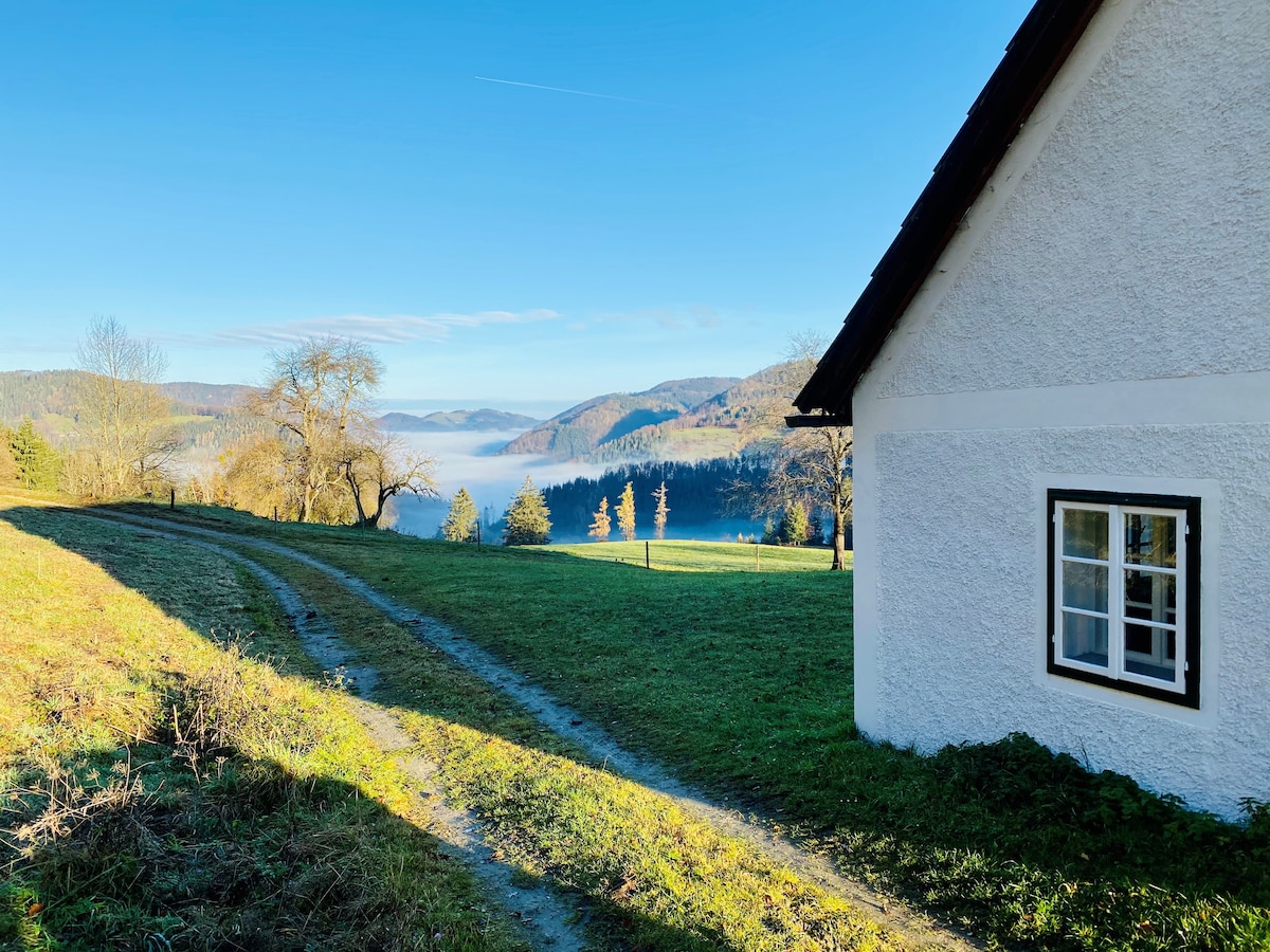 A scenic view of the countryside is visible from the edge of the property, with a gentle slope leading to lush green fields. A simple white house with a black roof is seen on the right, while rolling hills and mist-covered valleys fill the background under a clear blue sky.