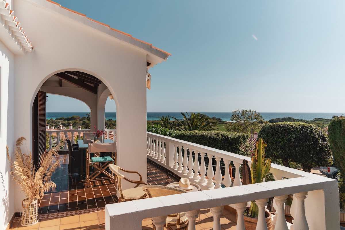 A spacious terrace is presented, overlooking the shimmering blue sea and lush greenery. A dining table is set for outdoor meals, accompanied by decorative plants. The architecture features white railings and tiled flooring, inviting a serene atmosphere enhanced by natural sunlight.