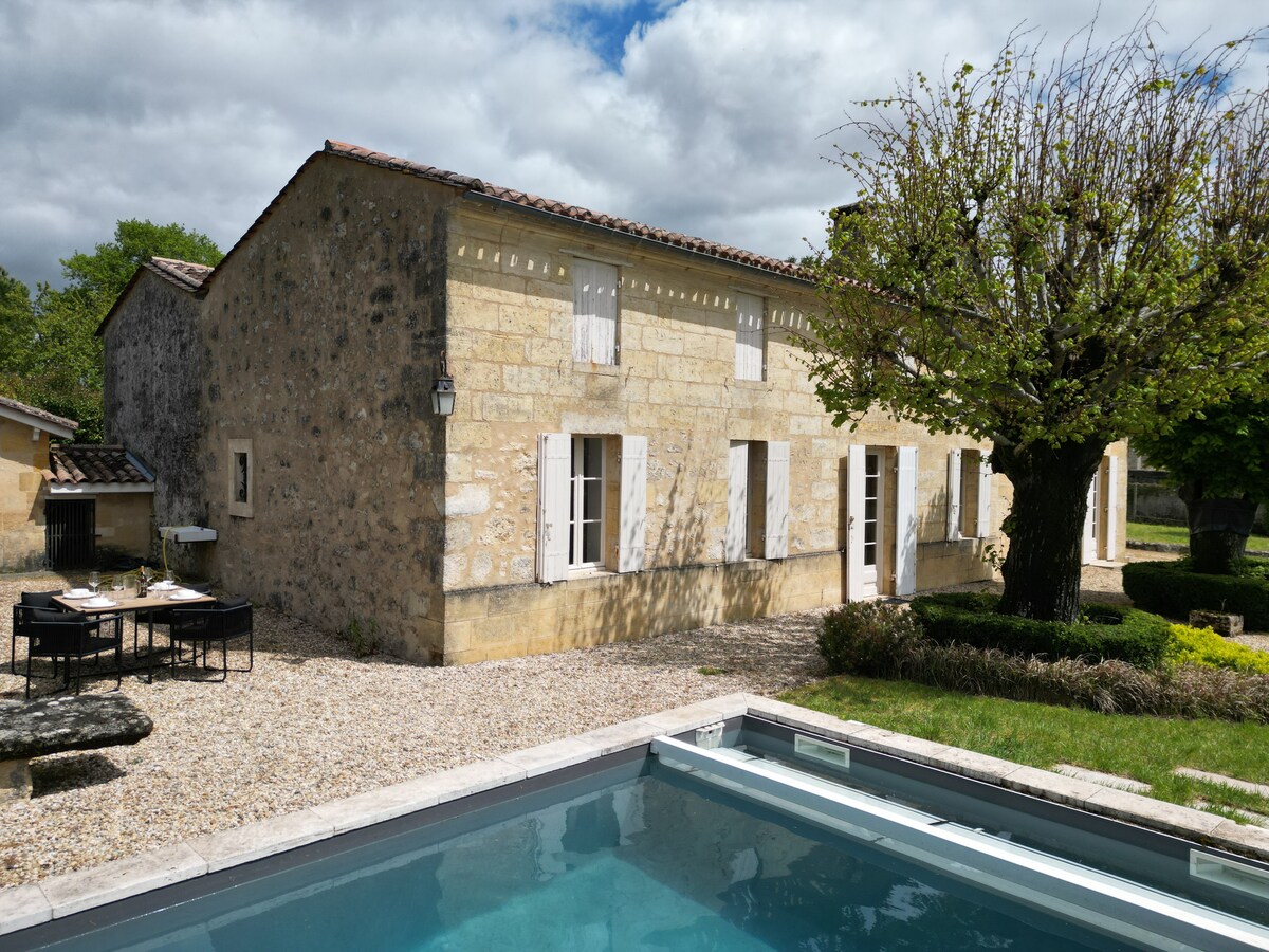 A rustic stone house is surrounded by a well-maintained outdoor space with a gravel pathway. A swimming pool sits at the forefront, complemented by a patio featuring a dining area. Lush greenery enhances the serene garden setting, while the sky is partly cloudy.