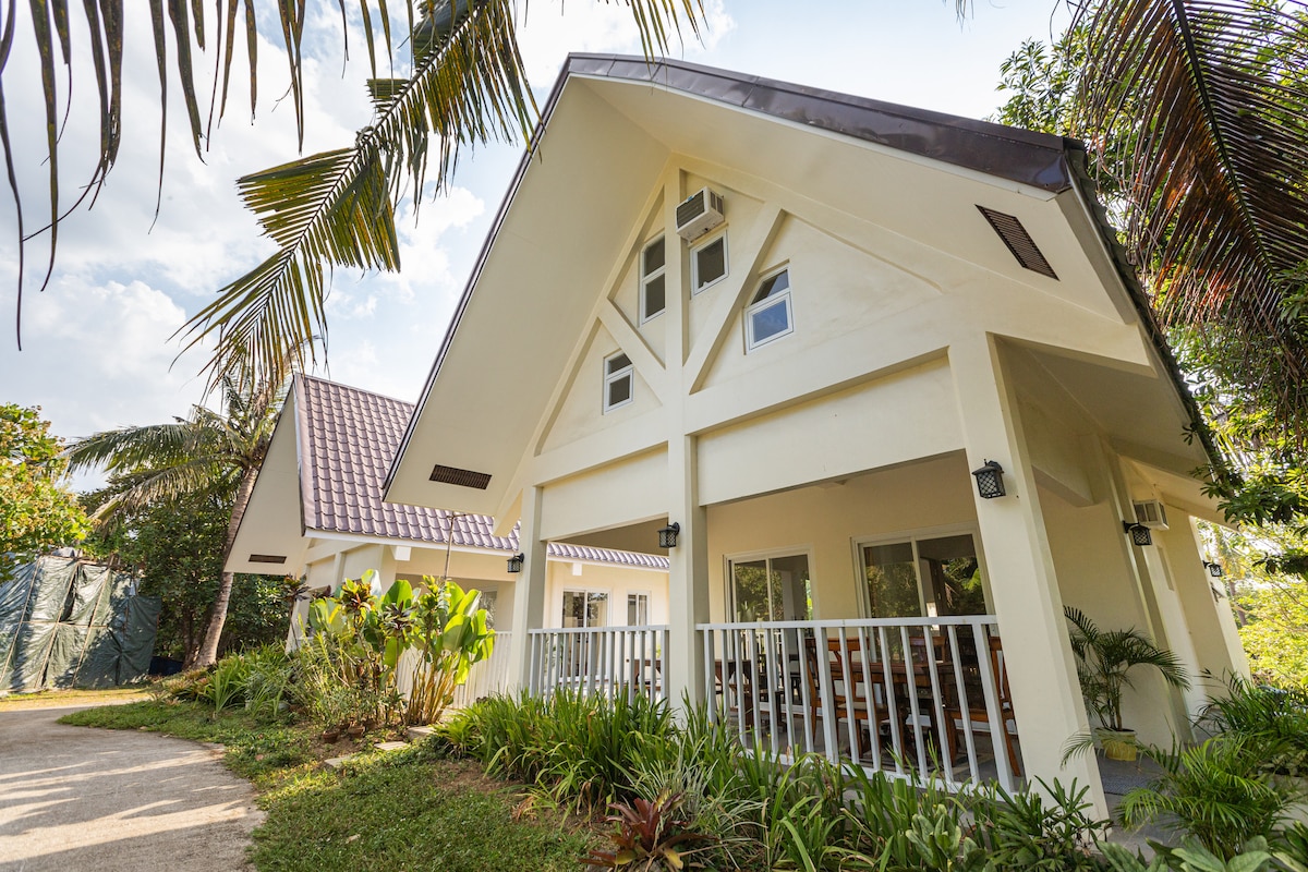 The twin farm homes are showcased with a sloped roof, surrounded by lush greenery. Large windows invite natural light into the living areas, while a covered porch provides access to the outdoor space. Tropical plants and palm trees dot the landscape, creating a serene setting.