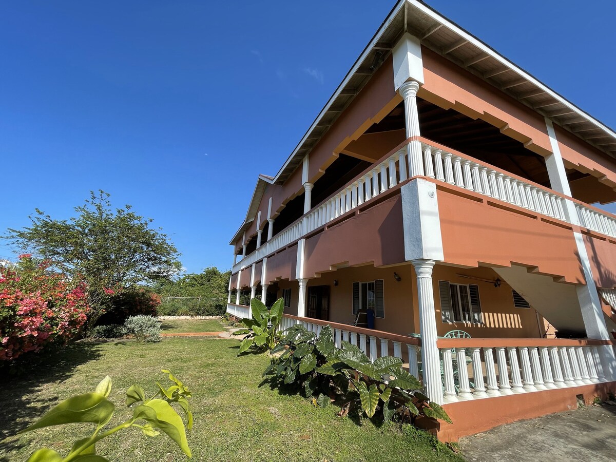 A two-story building with a warm orange exterior is surrounded by well-maintained greenery. Balconies with white railings provide outdoor spaces for relaxation. Sunlight filters through the trees, creating a serene atmosphere in the garden area.