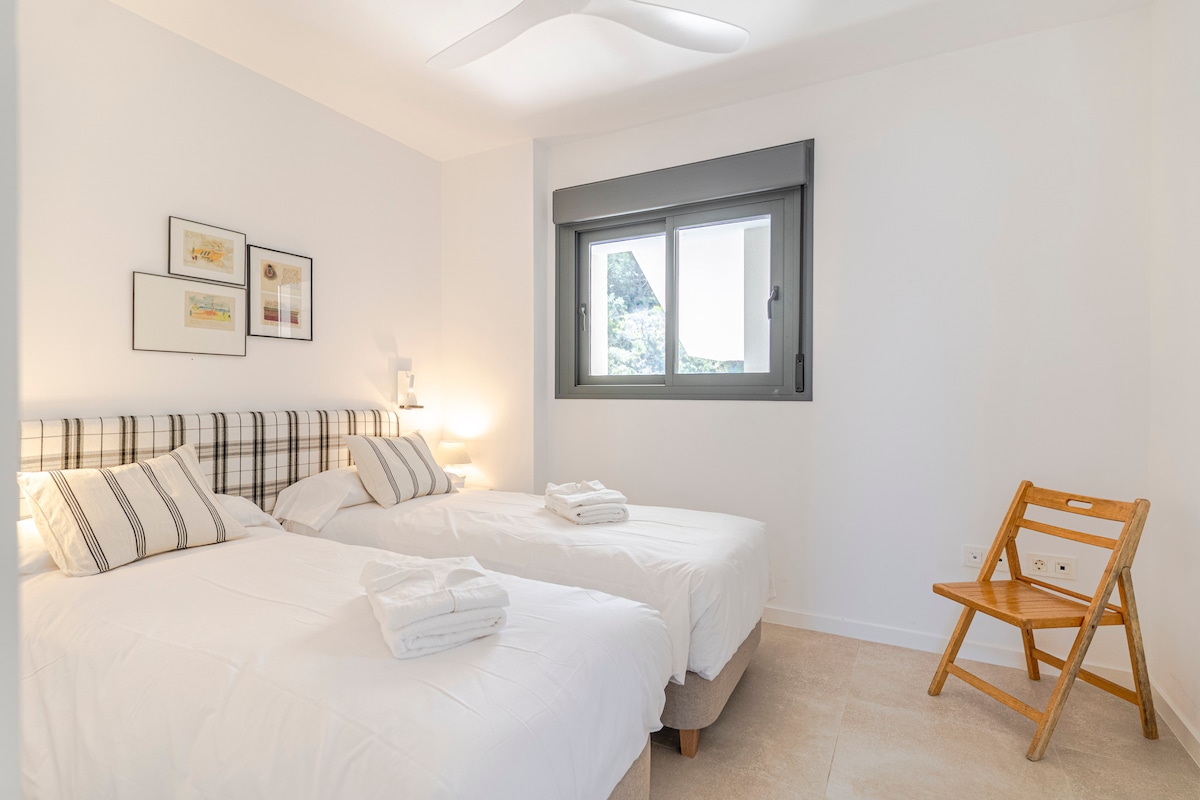 Dormitorio con dos camas individuales, cabecero acolchado, mesitas con lámparas, ventilador de techo, armario empotrado, silla plegable y ventana. Espacio funcional para dos personas, conectado por pasillo a baño y otras estancias del alojamiento.