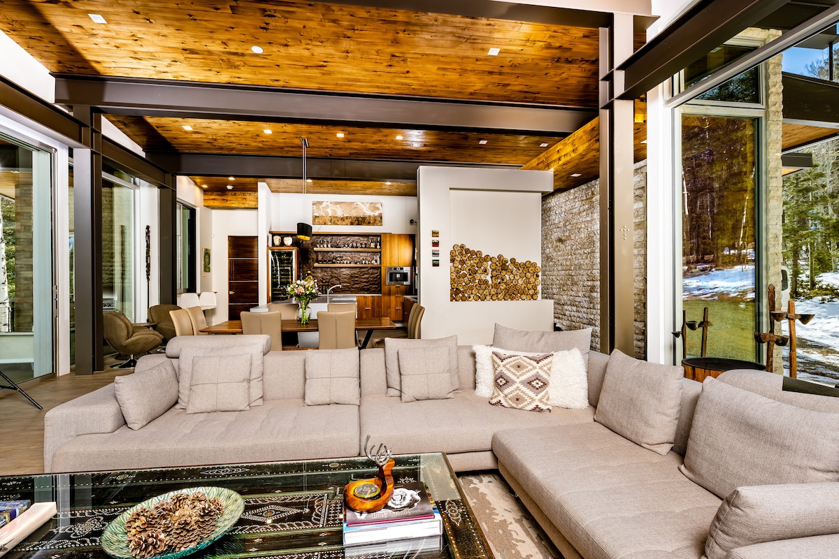 A contemporary living area features a spacious sectional sofa positioned around a low coffee table. Large glass doors provide access to the outdoor space, while the wood-paneled ceiling adds warmth. A dining area is visible in the background, complemented by artistic wall decor.