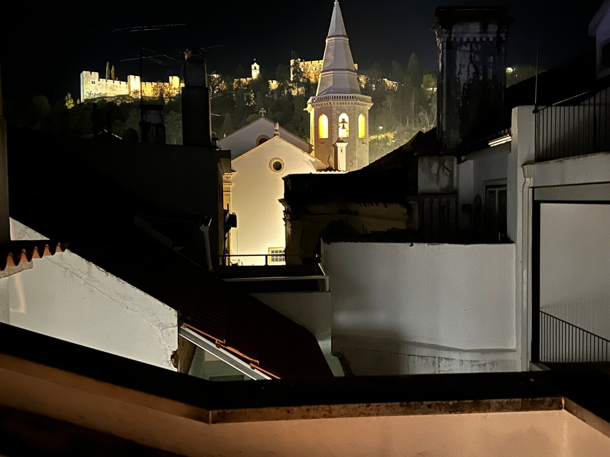 In the night scene, the rooftops of traditional buildings are visible, with varying heights and textures. A prominent steeple is illuminated, highlighting its architectural details. In the background, the castle stands on a hillside, softly lit against the dark sky.