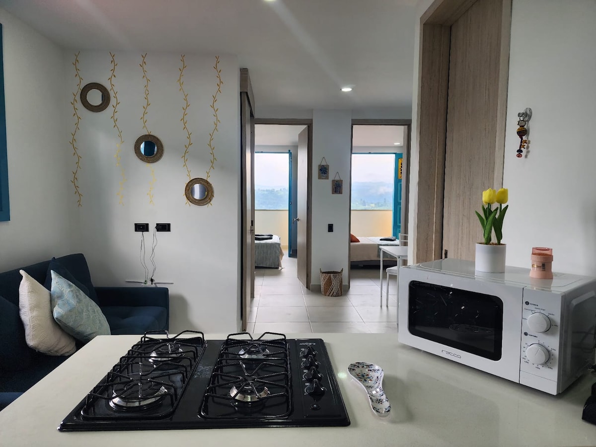 The image shows a modern kitchen area featuring a gas stove with multiple burners, a microwave, and small decorative items. The space is complemented by a comfortable seating area with a blue sofa and a glimpse of a well-lit hallway leading to the bedrooms, framed by large windows.