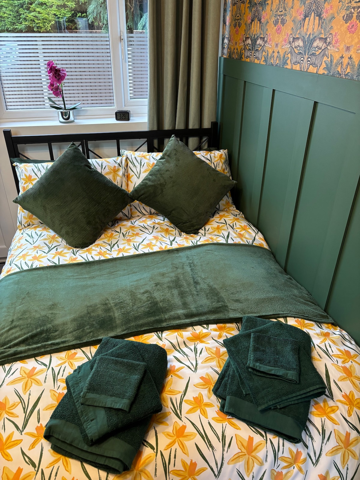 A well-prepared sleeping area features a double bed layered with floral bedding and an oversized green throw. Two decorative pillows are arranged at the head of the bed. Fresh towels in varying shades of green are neatly organized at the foot of the bed, enhancing the overall arrangement.