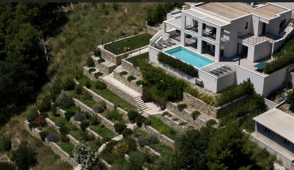An aerial view captures a modern villa surrounded by greenery, featuring a turquoise pool and spacious outdoor seating. Landscaped terraces with various plants and flowers lead downwards, while a grassy area offers additional recreation possibilities. The structure's white exterior complements the natural environment.
