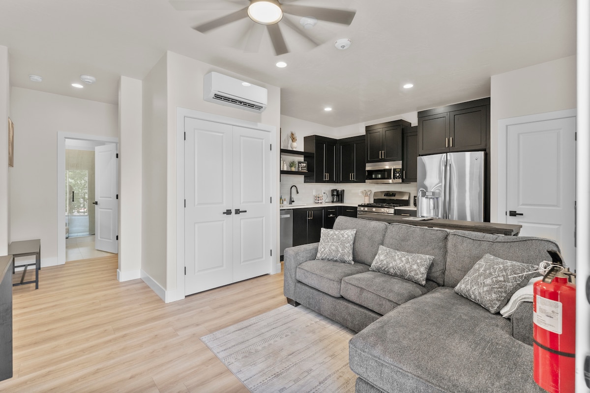 An open-plan living area showcases a modern kitchen with sleek black cabinets and stainless-steel appliances. A spacious gray sectional sofa is situated nearby, accompanied by decorative pillows. Light wood laminate flooring extends throughout the space, accentuating the welcoming ambience.