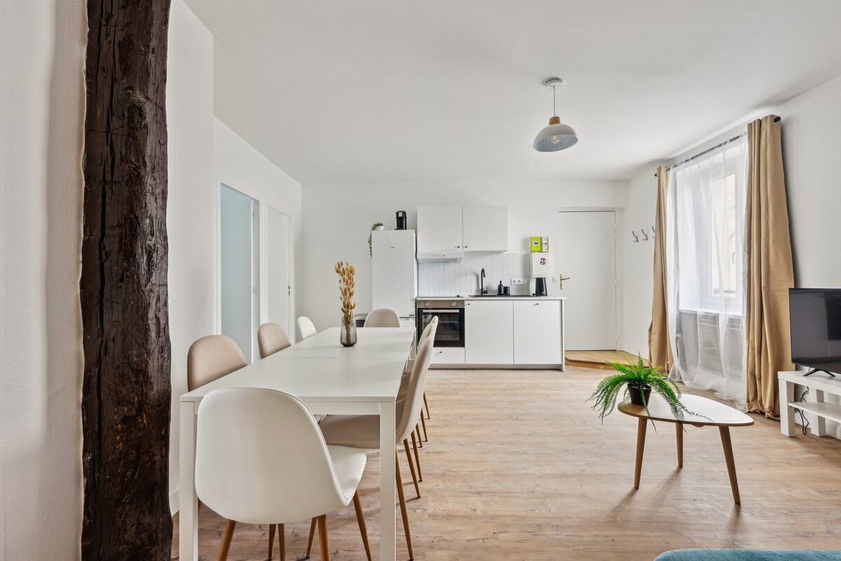 A bright and airy dining area is presented, featuring a white table surrounded by several chairs. The kitchen area is visible, showcasing modern appliances and cabinetry. Natural light streams in through large windows, and a small plant adds a touch of greenery.