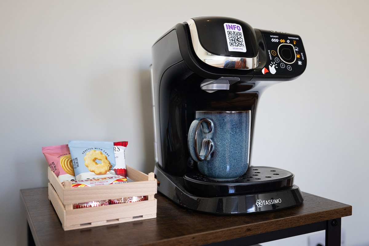 A coffee machine is positioned on a dark wooden table, with a filled cup placed underneath it. A small wooden tray holds an assortment of packaged snacks, providing convenient options for guests.