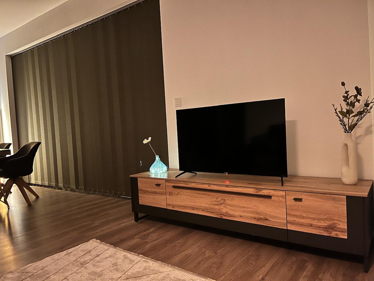 A modern living area features a wooden TV console with two drawers beneath a flat-screen television. Natural light filters through a vertical blind covering a large window, enhancing the warm tones of the laminate flooring. A decorative vase with a single flower adds a subtle touch of decor.